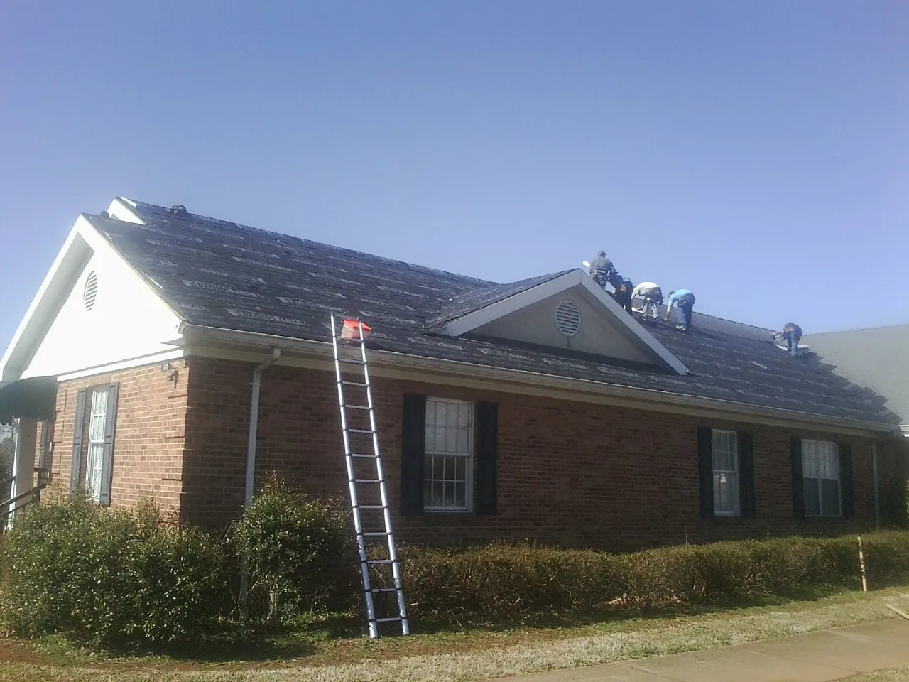 Crew tearing shingles off a brick ranch roof with a ladder set — Metal Roof Repair work by our crew in Pella