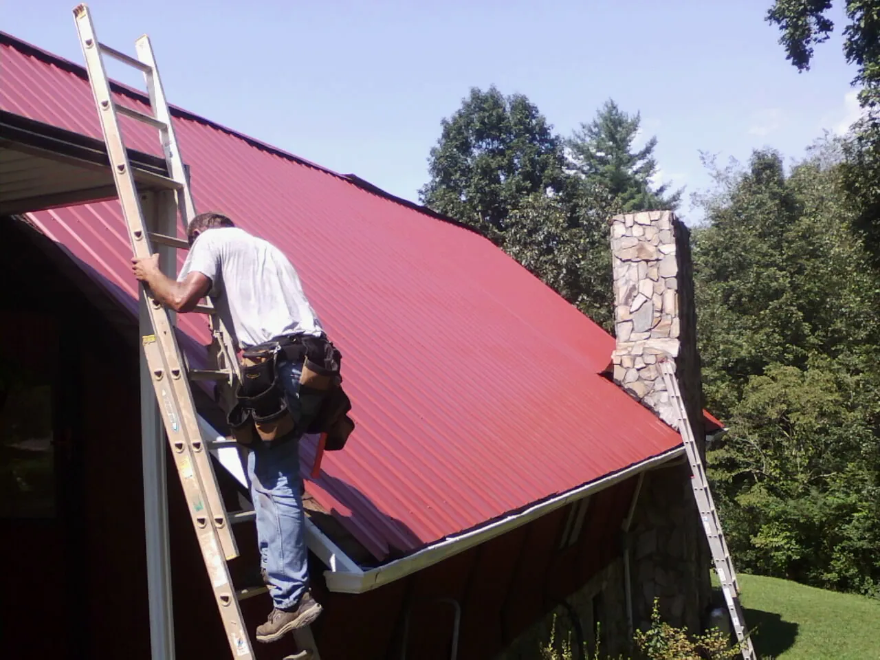 Roofer on a ladder beside a red metal roof and stone chimney — Soffit & Fascia Repair by Roofing Brothers in Pella